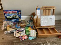 Full lot showing Rummikub game, acrylic paint sets, artist brushes, paint palette, canvas pad, and wooden easel with pull-out drawer.