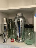 Silver-tone cocktail shakers and servers, double-sided jigger, stopper, and green-tinted blown glass wine glasses on white shelf.