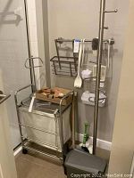 Wide view showing metal framed hamper with fabric bag, tall standing towel rack, mounted rail with hanging baskets, brushes, scraper, mirror, hooks, and caddies