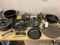 Photo showing the entire cookware set laid out on a wooden table including saucepans with lids, T-Fal stir fry pan with red heat spot, cast iron pans and a flat griddle pan.