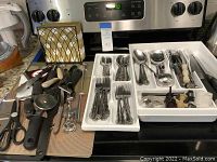 Full view of flatware organized in white trays and cooking utensils spread out on countertop showing variety and quantity.