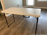White folding table fully set up with legs extended on wood floor.