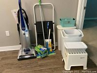 Full view of the lot showing vacuum cleaner, 2 step stool, plastic hampers, trash can, mop and cleaning supplies.