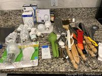 Wide view of assorted light bulbs and kitchen tools arranged on granite countertop, showing unopened boxes of GE Reveal and Ledare bulbs alongside various hand kitchen utensils and loose bulbs