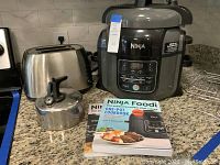 Ninja Foodi air fryer with instruction books and metal tea kettle on a kitchen counter.