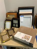 Various sizes and styles of picture frames including wood, black, gold-tone, and multisection designs arranged on table.