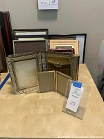 Assorted gold tone and wood picture frames arranged on table, including ornate embossed gold frames and plain black frames.