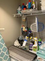 Photo of laundry and cleaning products on a wire shelf above an ironing board and some clothing items, also shows wire hat forms hanging on wall.