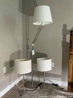 Full view of the set showing one tall silver tone adjustable floor lamp with white lampshade and two smaller white table lamps with textured cylindrical shades and square bases.