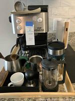 View of Via Venezia stainless steel espresso coffee maker with accessories including portafilter, tamper, milk pitcher, scoop, and French press on kitchen countertop.