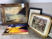 Four framed artworks displayed together including a large oil painting with gold ornate frame, a needlework picture, an Asian ink drawing, and a colorful beach scene painting