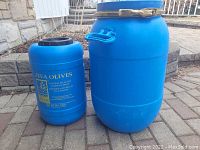 Two blue plastic barrels placed outdoors on stone tiling for size and condition display. Larger barrel has lid and handles, smaller one has printed branding.