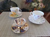 Three coffee cups with saucers displayed on a mat with floral painting background. Cups have distinct floral and decorative patterns.