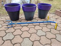 Photo shows three purple-rimmed gray plastic patio containers, approximately 15 inches tall, arranged in a row outdoors on a patterned stone surface. In front lies a blue garden cultivator tool with yellow grips on the handle end and three metal prongs on the other end.
