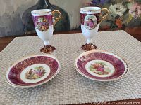 Two porcelain footed cups and two saucers displayed on a table with decorative artwork and gold accents.