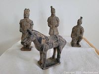Four terracotta-style figurines on white cloth backdrop, showing three warrior statues and one horse statue from front angle.