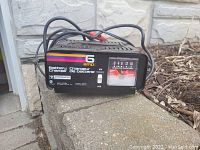 Motomaster 6 Amp battery charger shown on outdoor stone ledge, featuring voltage selector and analog amp meter with cables attached.