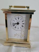 Front view of Bulova clock showing gold-tone rectangular frame, white dial with Roman numerals and decorative floral etching below clock face.