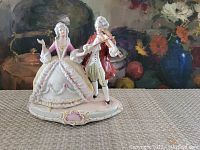 Full view of porcelain figurine showing an 18th-century couple with the man playing a violin and the woman posed with her arm extended. The base has pink and gold decorative accents.