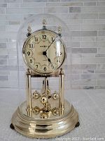 Close-up of a Howard Miller anniversary clock with brass-tone base, clear glass dome, cream clock face with floral details and black numerals and hands.