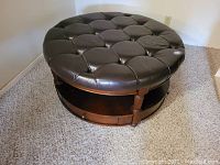 Front angle of round leather coffee table showing tufted leather top and wooden base with open space below.