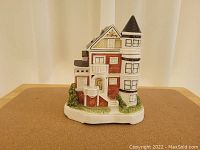 Front and side view of a ceramic musical bank shaped like a Victorian house with turret and detailed paintwork.