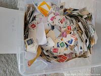Top view of a plastic container filled with assorted old postage stamps affixed to paper pieces, showing many colors and designs.