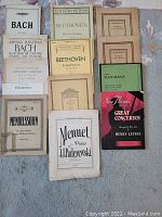 Photo of thirteen piano sheet music and books laid out on fabric surface, showing titles and cover designs.