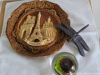 Photo showing the Paris souvenir plate, cast iron dragonfly paper holder, and glass paperweight displayed together on a white cloth.