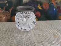 Clear crystal candle holder with rock cut geometric pattern on all sides, displayed on a textured beige surface with floral artwork background.