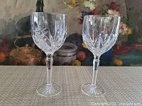 Two clear Waterford Marquis crystal goblets side by side on a woven textile surface with floral painting background