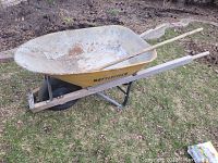 Full view of the yellow Battlefield wheelbarrow with a single rubber tire and wooden handles, resting on grass near a garden bed.