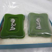 Two miniature pewter figures mounted on green velvet cushions with decorative edging, displayed side by side in a box.