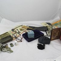 Wide shot of assorted vintage and antique items laid out on a white cloth including keys, glasses, coins, silverware, photos, and small boxes.
