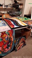 Seven oil paintings laid out on tables and leaning against furniture. Various sizes and styles visible, vibrant colors and patterns.