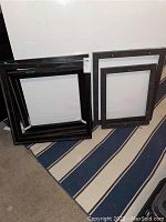 Photo showing multiple black wood frames of different sizes leaning against a wall on striped rug.