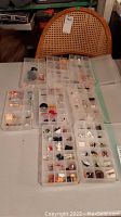 Eight clear plastic storage containers with multiple compartments, each housing various miniature figures and items, spread out on a table with a chair in the background.