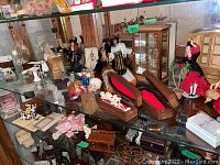 Glass display case with miniature Halloween figures including skeletons in wooden coffins, witches, dolls in costumes, skulls, and miniature furniture such as cabinets and tables.