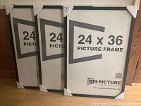 Three new 24x36 inch picture frames stacked leaning against a wall, each protected with corner cardboard covers, showing the Big Picture Custom Framing brand on paper insert.