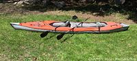 Side profile of fully inflated orange and gray kayak on grass with two black paddles attached