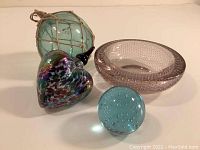 Four colorful art glass pieces displayed together on a table: green fishing float, pink bowl, blue globe, and multicolored heart ornament.