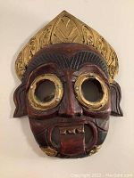 Front view of Balinese carved wood Rakshasa Bhoma mask with gold and black paint detailing and circular eye holes.