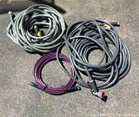 Three garden hoses coiled on ground: gray (large and medium size) and purple (small size) hose, showing various ends and attachments.
