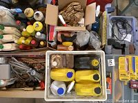 Top-down view of boxed assorted bottles and containers of chemicals and liquids, including yellow and white jugs.
