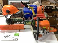 Full lot overview on tabletop showing chainsaw, bar cover, instruction manual, helmet, earmuffs, fuel containers, oil bottle