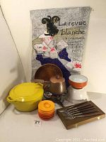 Photo showing the items: yellow Copco D3 dutch oven with lid, copper fondue pot with stand and handle, white mixing bowl with orange rim, farm design platter, Copco enamel bowl, set of colorful egg cups, and French tea towel with vintage artwork hanging behind