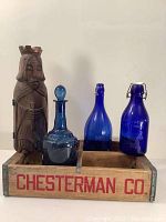 Front view of lot showing carved wooden king figure wine case, two blue glass milk bottles, one blue glass decanter, and wooden Chesterman crate.