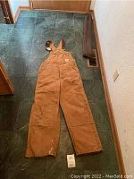 Full view of brown Carhartt men's work overalls laid flat on dark tile floor showing front side with bib and legs.