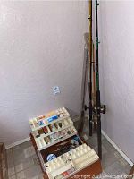 Full view of the fishing rods standing in a corner with a wooden tackle box open on the floor with multiple compartments containing fishing lures and other gear.