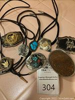 Overview photo showing multiple men's belt buckles and bolo ties arranged on surface with varied finishes including silver, copper, turquoise, and gold tones.